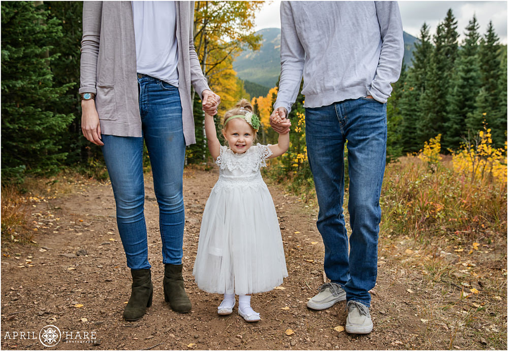 A cute little girl with blonde hair wearing a white dress holds hands with her parents at their outdoor fall color family session