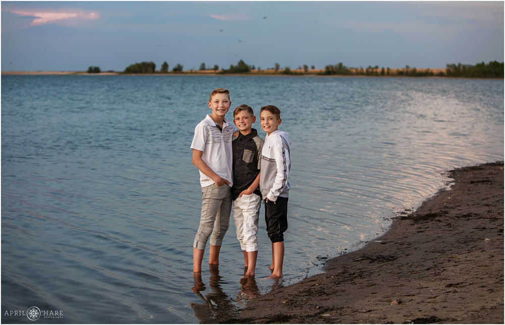 3 brothers pose for a portrait as they stand barefoot in the pretty blue water of Aurora Reservoir at Sunset