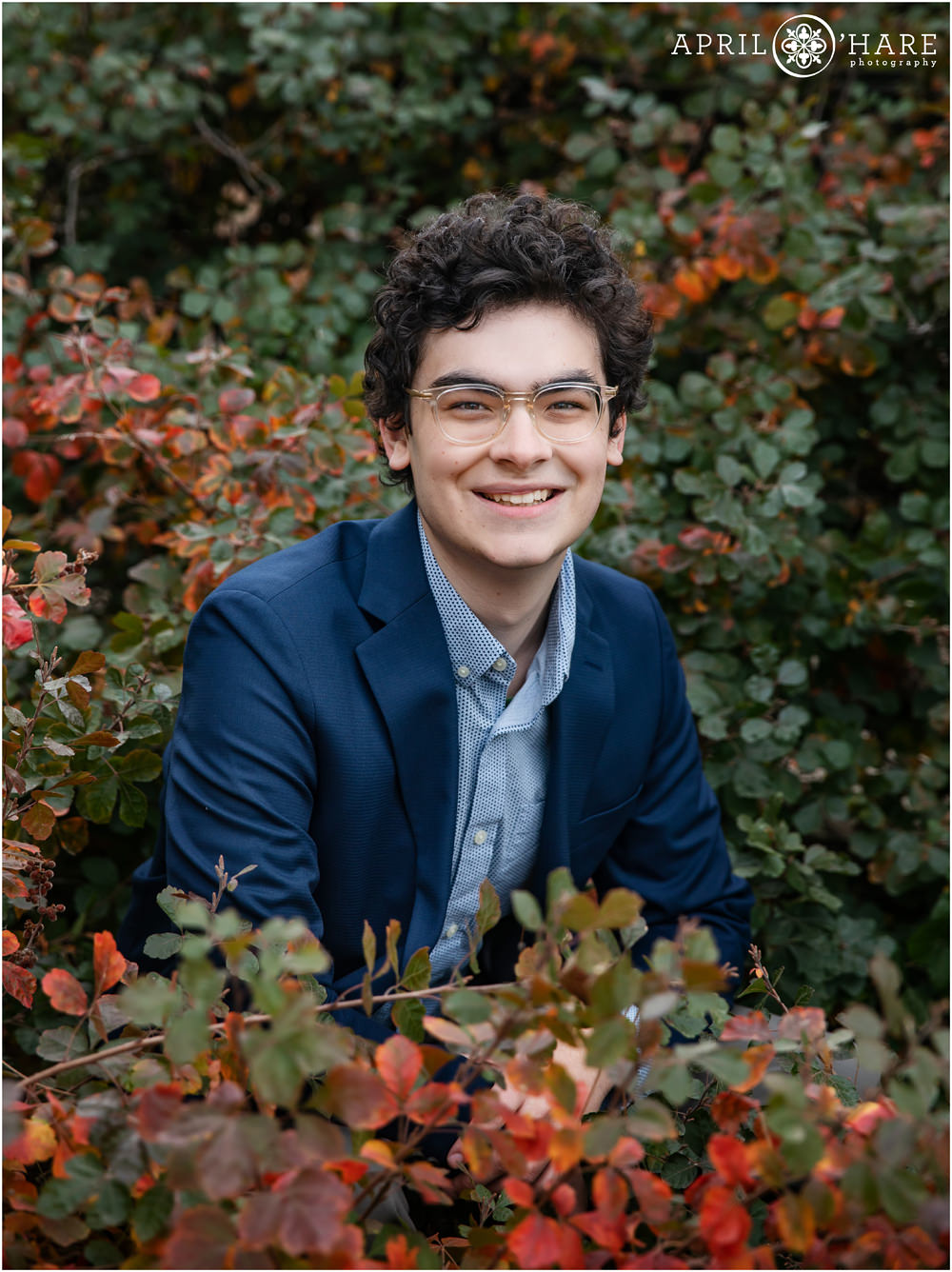 Beautiful fall color foliage provides scenery for a senior boy with dark curly hair and glasses to pose in at his downtown Denver senior photo session
