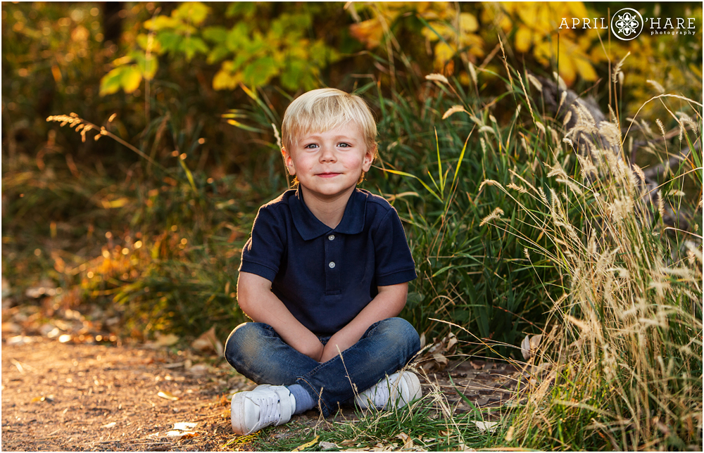 Cute little three year old blonde boy wearing blue polo shirt and blue jeans sits on a path in the fall color at the Mary Carter Greenway Trail in Littleton Colorado