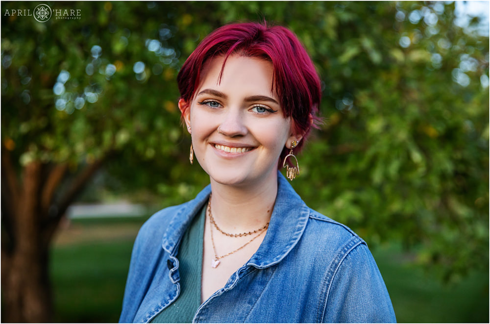 Beautiful high school senior girl with bright magenta colored hair with green tree leaves backdrop at City Park in Denver