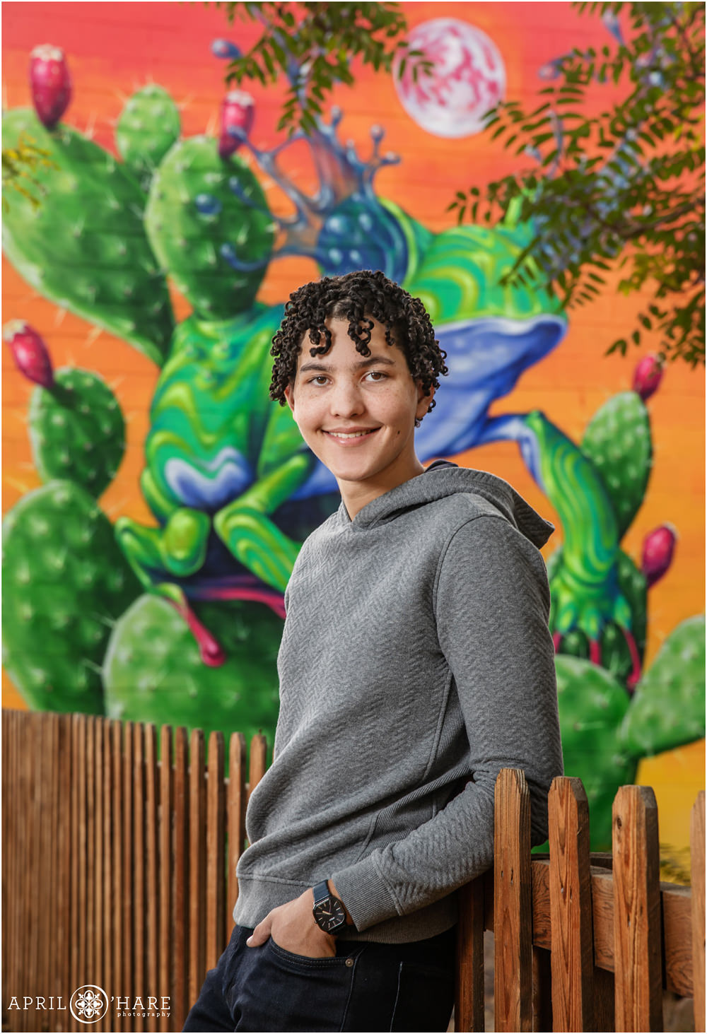 High school senior boy wearing a gray sweater, jeans, and a watch poses in front of a brightly colored frog and cactus mural in Five Points Denver CO