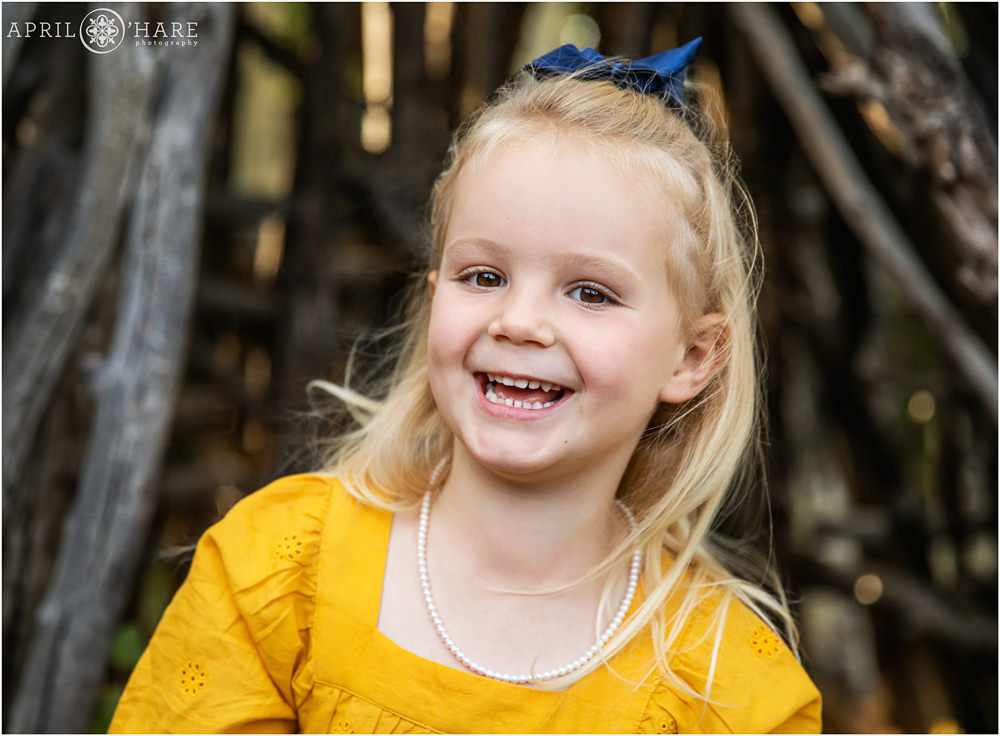 3 year old smiles big while wearing a blue ribbon in her hair and a bright yellow dress at her fall color family photos