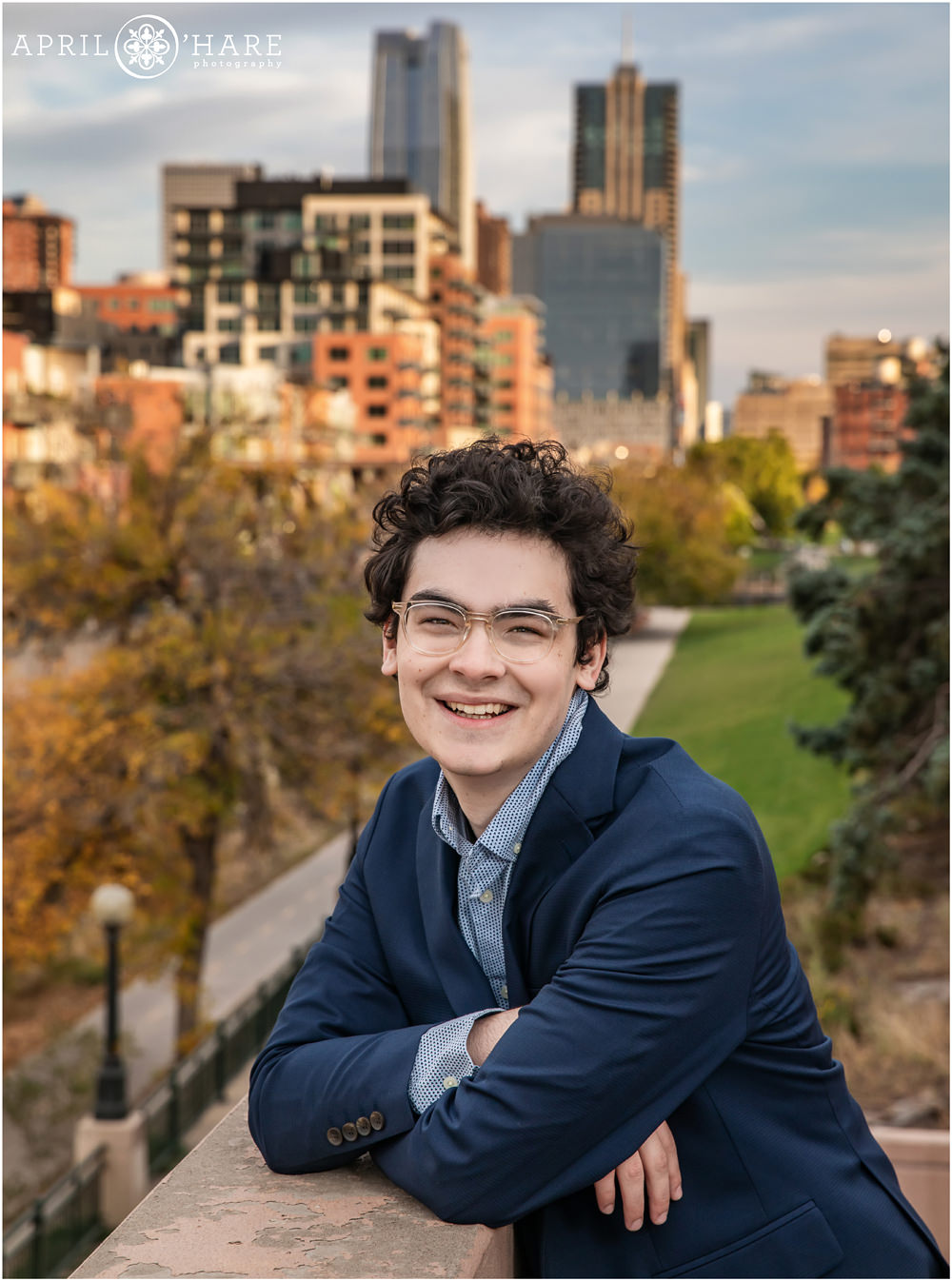 Young man with dark curly hair and glasses poses on a staircase along the Cherry Creek Trail in downtown Denver with a view of the city in the backdrop