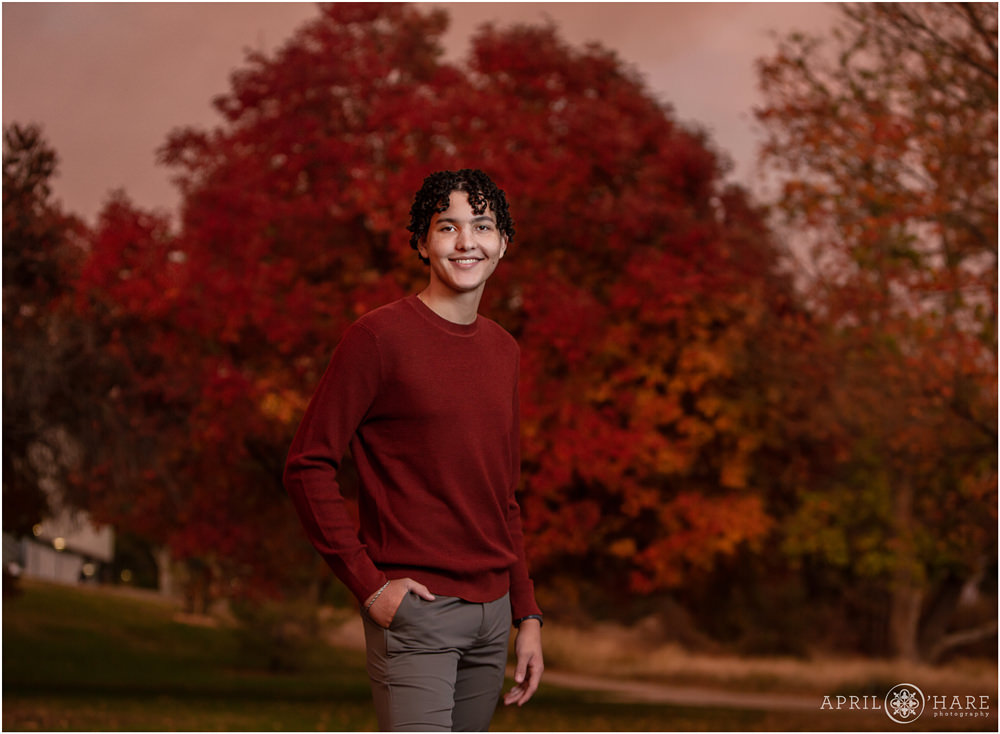 A senior boy wearing a burnt orange red sweater matches the fall color of a tree behind him on a smoky evening at City Park in Denver Colorado