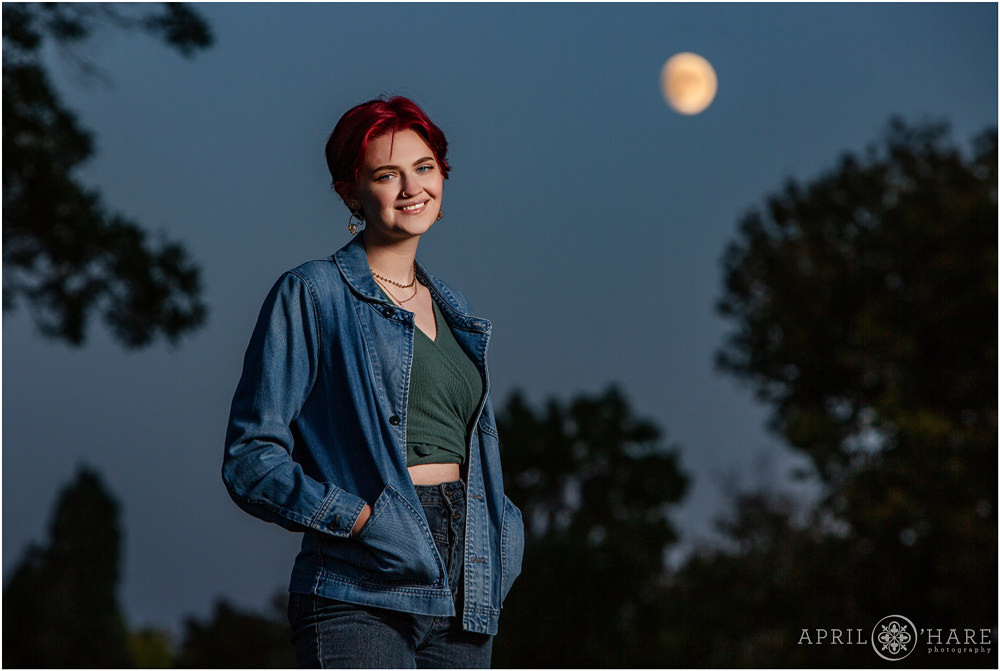 Beautiful high school girl with short bright red hair is photographed at dusk with a full moon backdrop at City Park in Denver Colorado