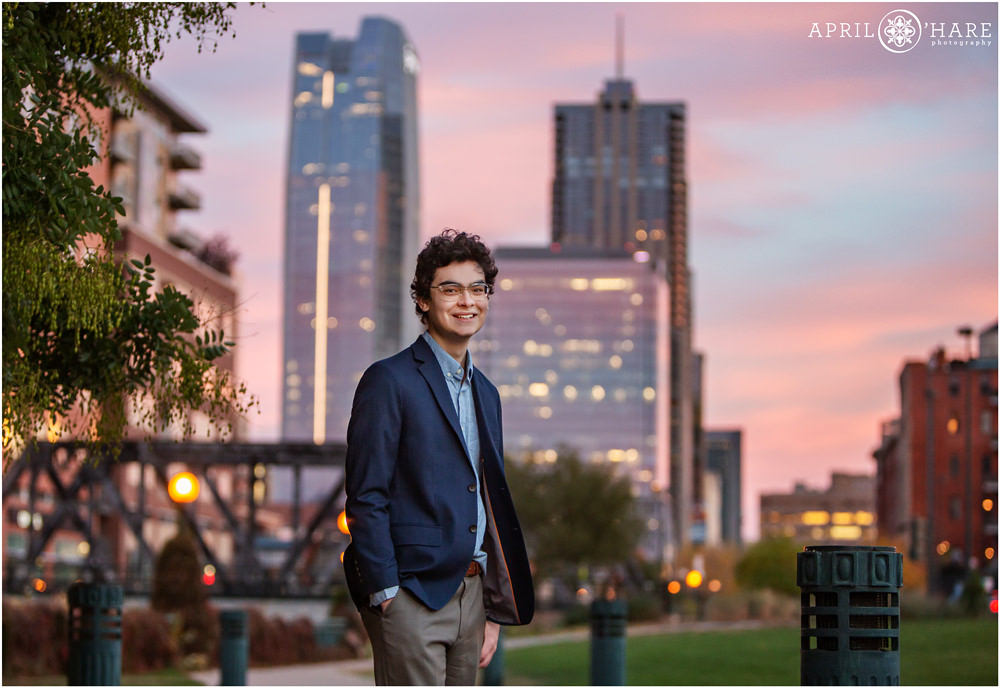 High School Senior Boy poses for a portrait wearing a dark blue suit jacket with Denver Skyscraper buildings in the backdrop at Sunset