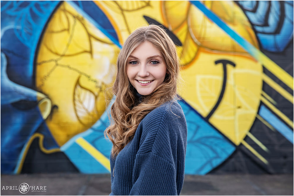 Pretty senior girl with long curled hair poses in front of a blue and yellow smiley face mural in North Denver