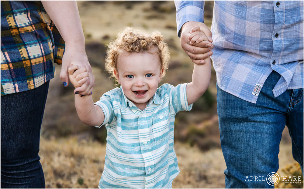 Sweet little boy with adorable curly blonde hair smiles big as he holds his parents hands at the East Mount Falcon Trail in Morrison Colorado