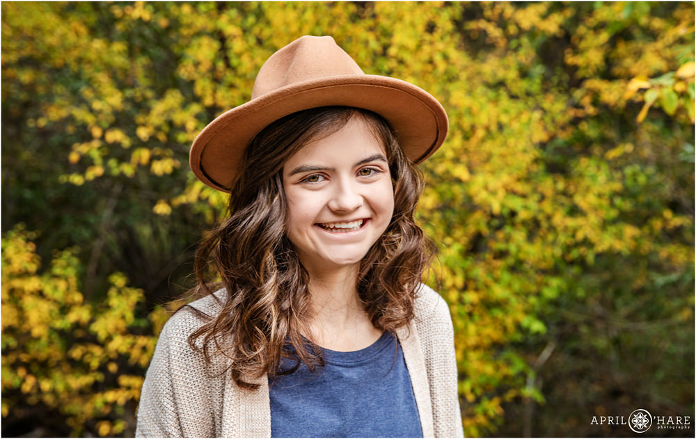 Pretty teen girl with curly brown hair wearing a cute wide brimmed hat poses in front of a fall color backdrop at her family portrait session in Estes Park