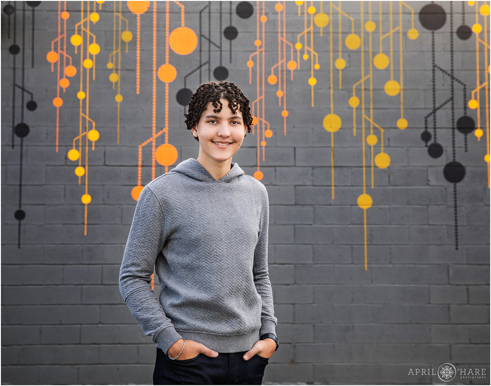 Senior boy wearing a gray sweater poses in front of a gray brick wall with orange and black geometric mural by Sandra Fettingis in Denver CO