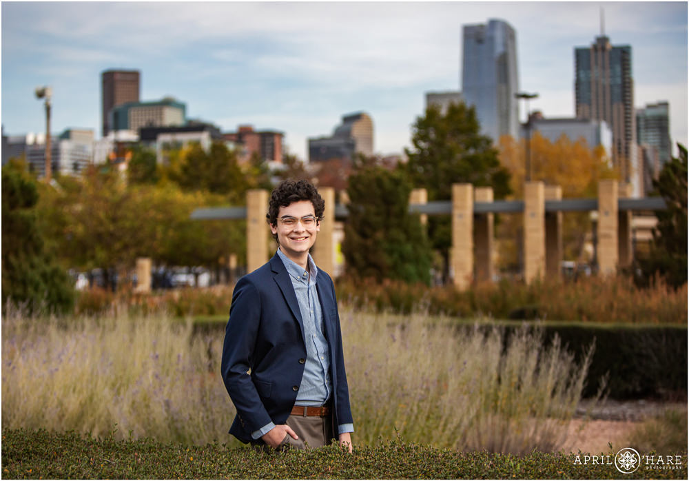 Denver senior boy with dark curly hair wearing glasses and a dark blue suit jacket poses at Centennial Gardens with a view of the City of Denver in the backdrop