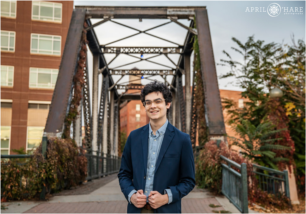 A senior boy with dark curly hair and glasses wearing a dark blue suit jacket poses with a cool old iron railroad bridge in the backdrop in downtown Denver CO