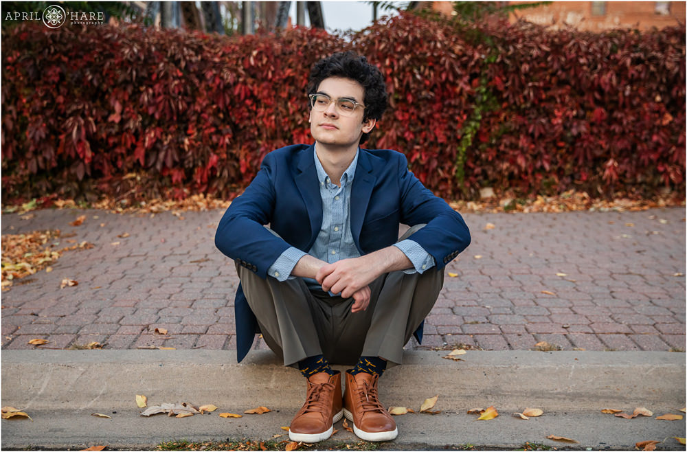 Fall color ivy wall backdrop for a young senior boy wearing glasses, dark blue suit jacket, and brown leather shoes in downtown Denver Colorado