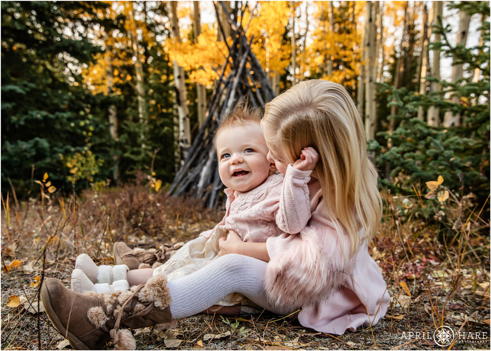 Two young sisters sit on the forest floor together and one gives her baby sister a kiss
