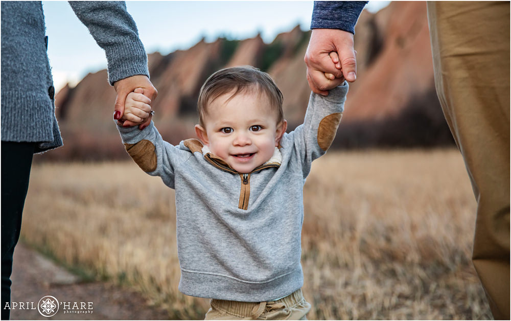 A sweet smiling toddler boy holds hands with his mom and dad with the pretty red rock formations in the backdrop at Roxborough State Park
