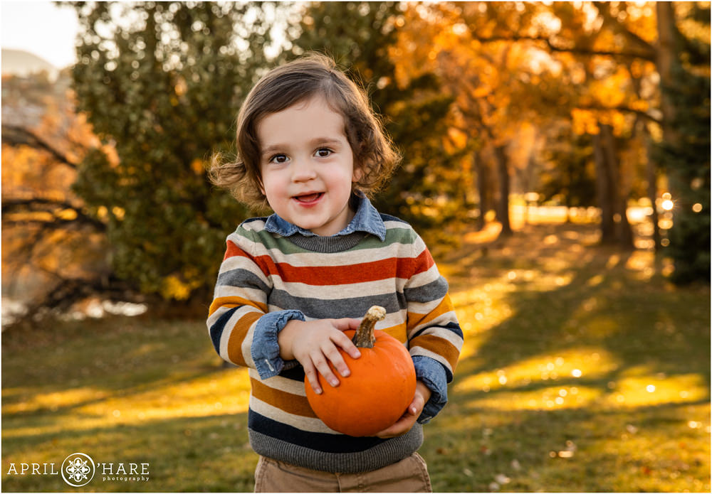 Sweet little boy wearing a fall color striped sweather holding a mini pumpkin in the fall color at Viele Lake in Boulder Colorado