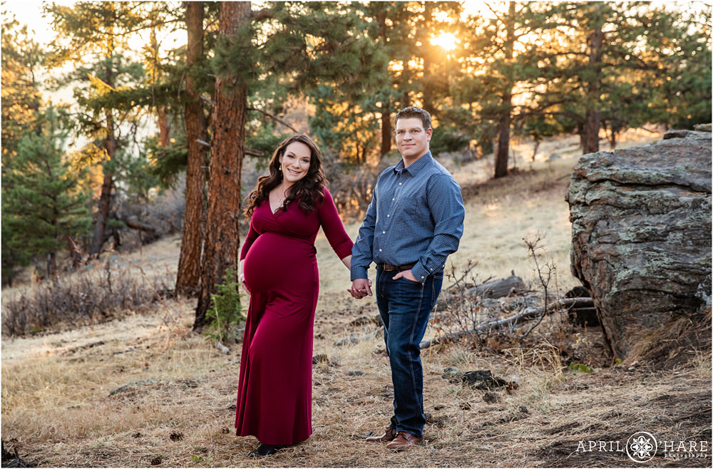 Gorgeous maternity photo with mama to be in floor length burgundy gown holds hands with her husband with pretty sunflare background