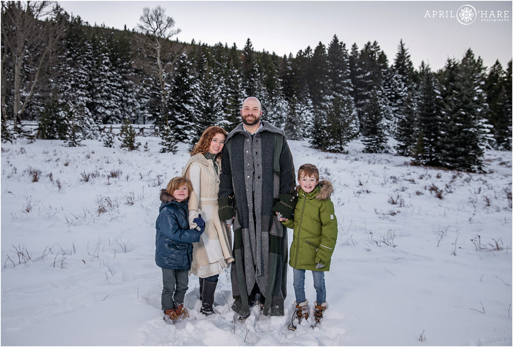 A family of four pose together with a pretty winter wonderland background full of snow covered evergreen trees