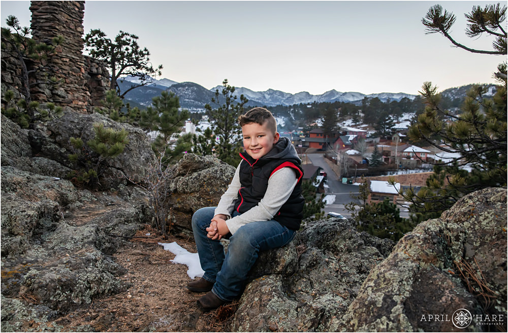 A 7 year old boy wearing jeans and boots sits on a rock near the Knolls-Willow Open Space Ruins with the town of Estes Park and mountains in the backdrop