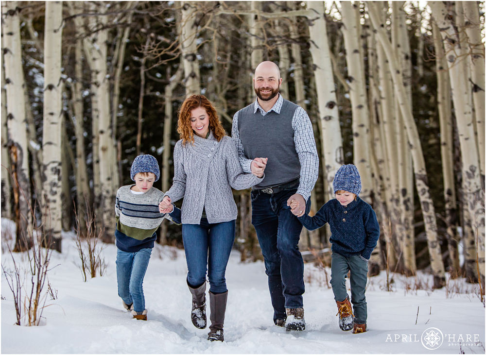 A cute family of four wearing shades of blue, green, and gray walk along a snowy forest path with aspen tree backdrop during winter