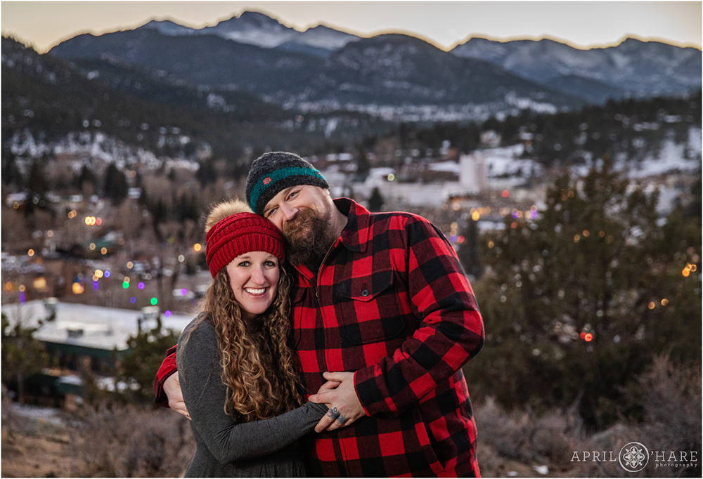 A couple wearing red plaid and winter hats poses in front of the pretty lights of the town of Estes Park during the holiday season