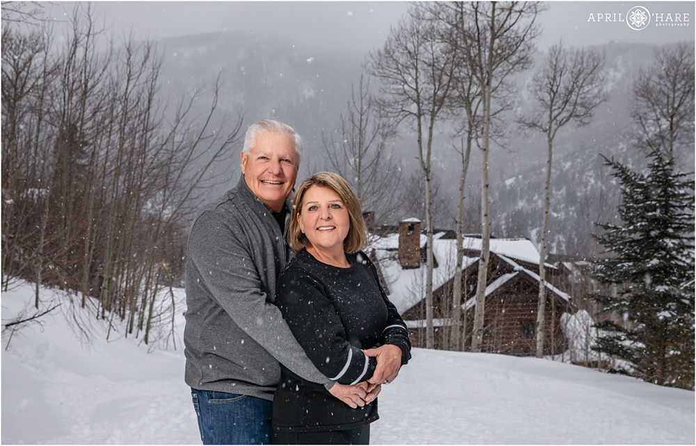 Couple about to celebrate their 50th wedding anniversary pose for a photo with a mountain backdrop on the catwalk outside of their vacation home in Beaver Creek