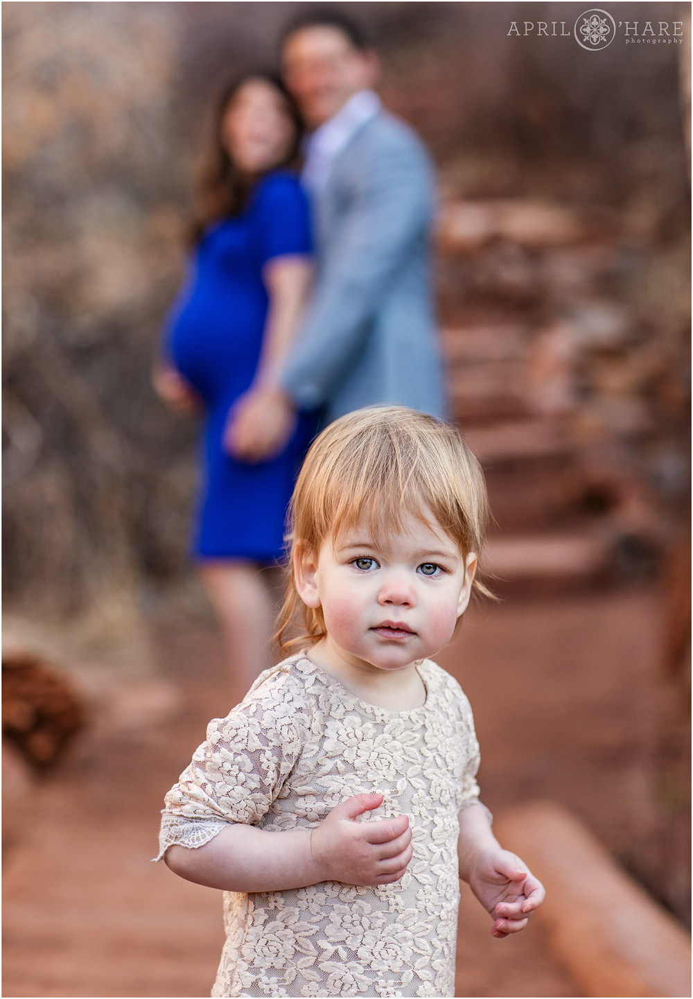 Cute little Girl wearing a light pink lace dress in the foreground with her pregnant mama and dad in the backdrop at Red Rocks Trading Post Trail in Colorado