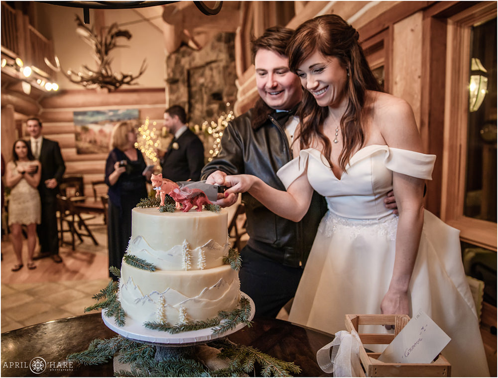 Brunette bride wearing off the shoulder romantic dress cuts cake with her groom wearing a flight jacket at their VRBO rental in Keystone CO