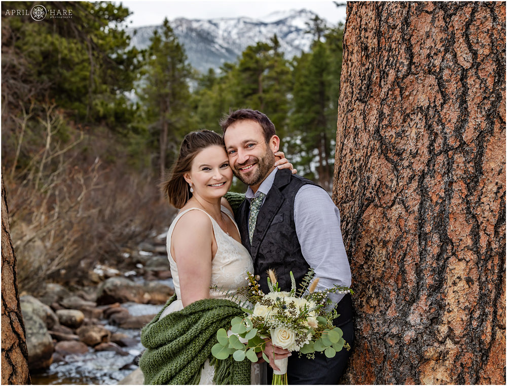 Bride and groom with big smiles pose next to large tree with Big Thompson River and Mountain Backdrop on their elopement day at Rocky Mountain National Park in Colorado