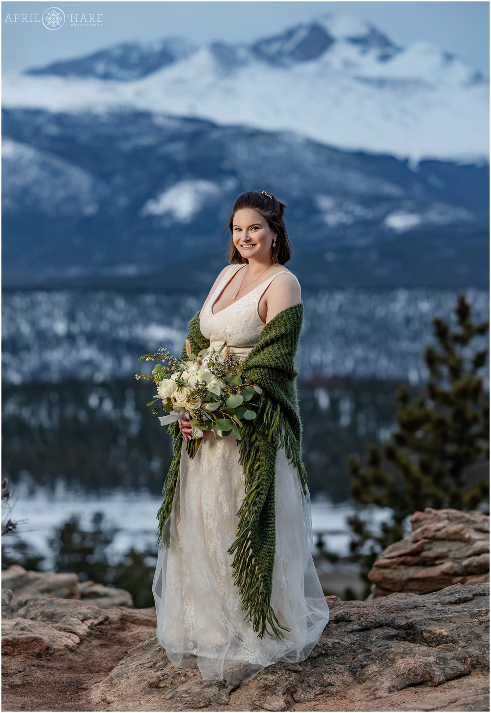 Full length bridal portrait for a pretty bride with short brunette hair holding a nice green and white bouquet and wearing a champagne colored gown with a green cozy shawl standing at 3M Curve with mountain backdrop