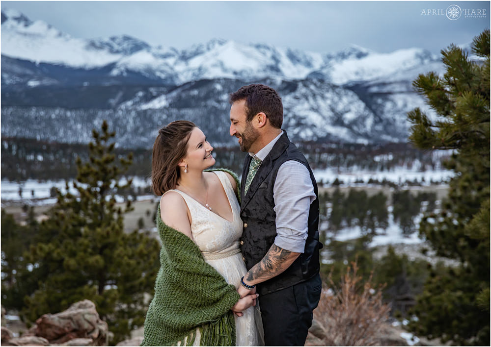 Sweet candid picture of a bride and groom smiling at each other in front of a beautiful blue mountain backdrop with lots of snow on it at 3M Curve in Estes Park