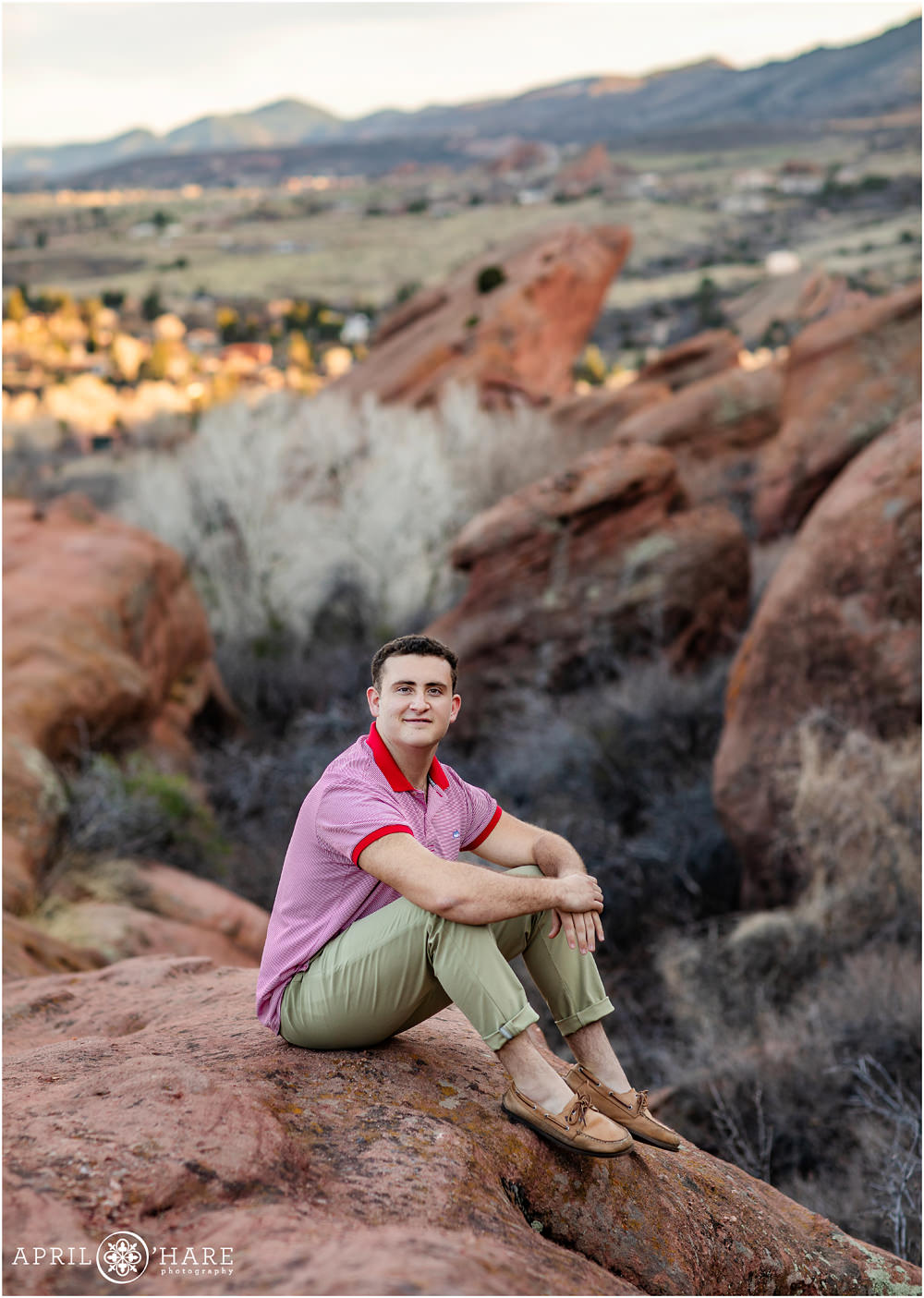 Teen boy wearing a red and pink shirt sits on the rocks at Red Rocks for his senior portraits in Colorado