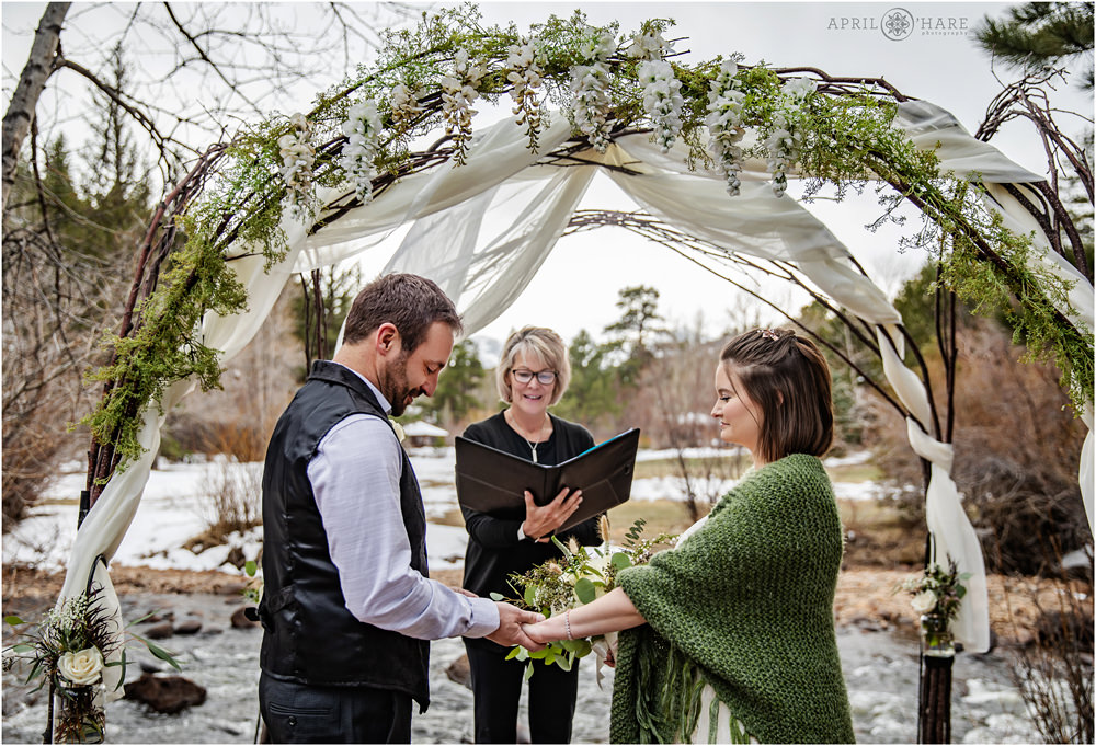 Groom puts ring on his bride's finger at their outdoor spring elopement at the Romantic RiverSong Inn next to Big Thompson River in Estes Park