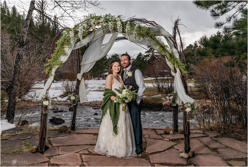 Formal posed wedding portrait of a couple standing under a beautiful wood arch decorate with white fabric and green and white florals next to Big Thompson River at Romantic RiverSong Inn in Estes Park CO