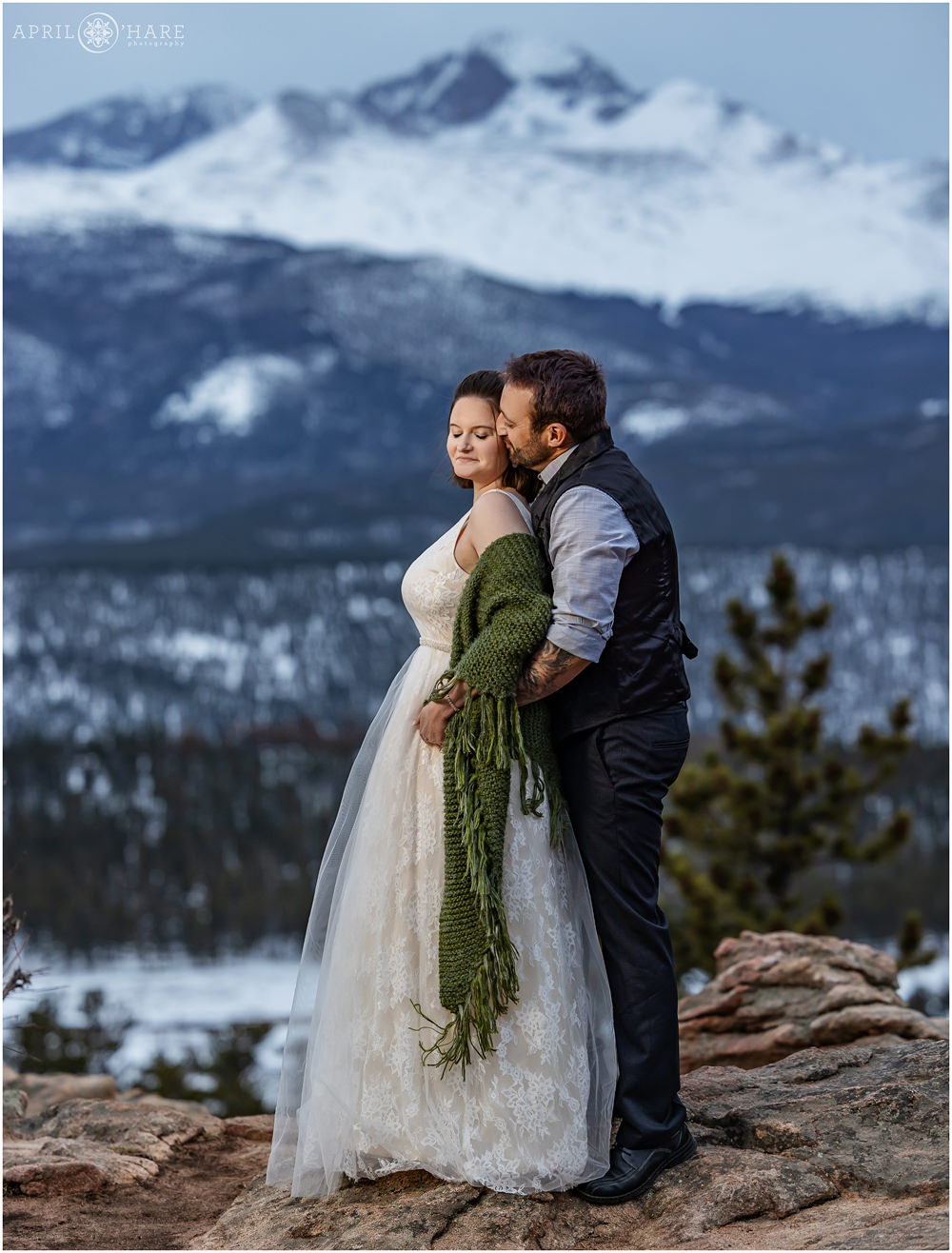 Groom snuggles into his bride as they stand on a rocky outcrop at 3M curve with a pretty blue mountain with lots of snow on it on their Spring Elopement day in Estes Park