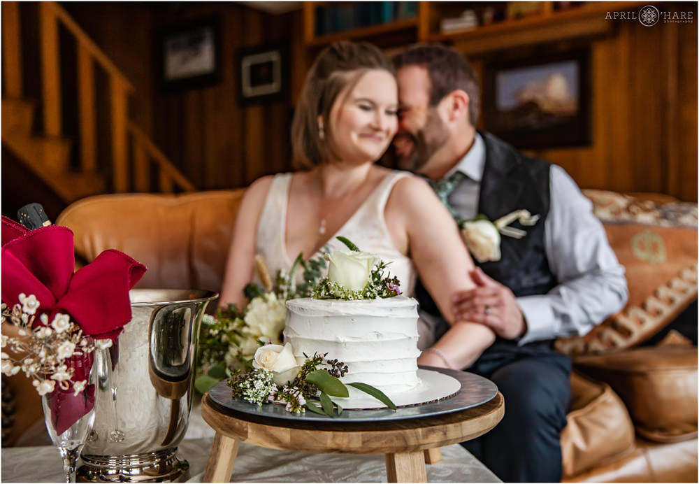 Groom snuggles his bride on the couch inside the Romantic RiverSong Inn with their cute white cake in the foreground with green and white florals in Estes Park