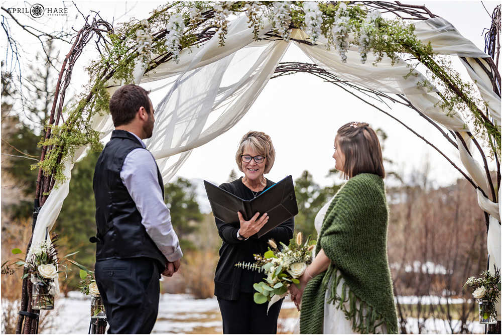 Cheryl from Romantic RiverSong Inn officiates a spring elopement wedding in Estes Park at their outdoor arch covered in green and white florals