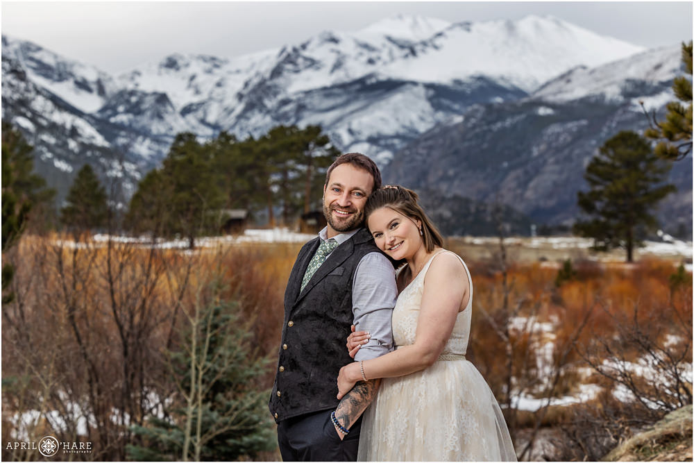 Groom wearing a chambray blue shirt with green tie and black vest poses in front of a beautiful snow covered mountain view as his pretty bride with short brunette hair snuggles into him at Rocky Mountain National Park