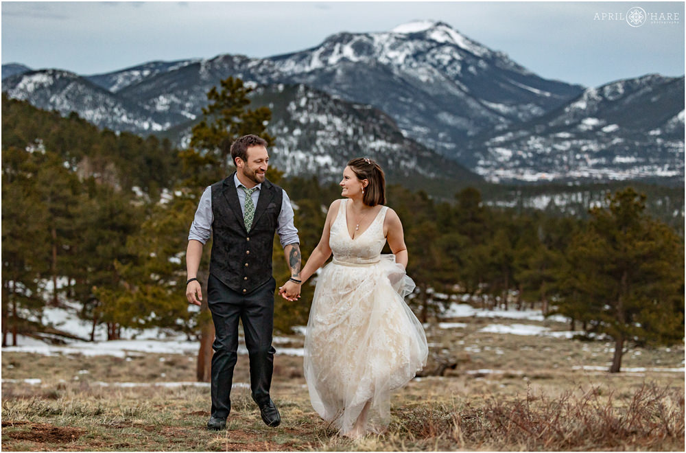 Bride & Groom hold hands while walking together as they smile at each other during spring at Rocky Mountain National Park with snow on the mountain