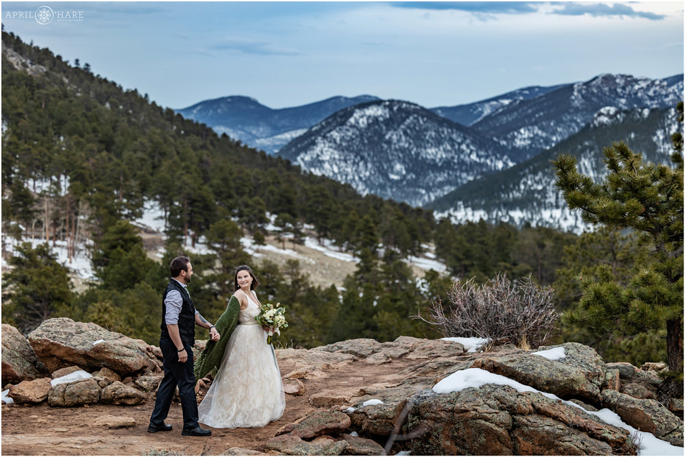 Couple walk hand in hand to the 3M Curve rocky overlook with a mountain backdrop with bits of snow on the ground during spring in Estes Park