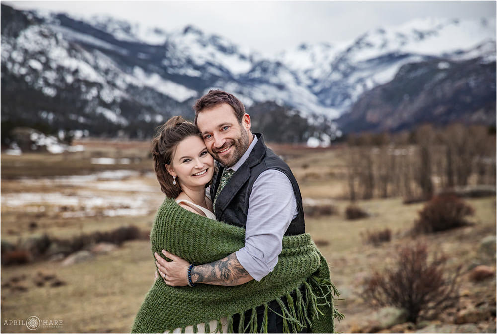 Bride with bob hair cut with a sage green sweater shawl snuggles her new husband at their elopement with mountains of Moraine Park in the backdrop on a spring day in Estes Park