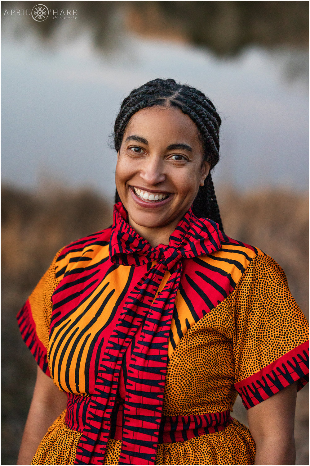 Beautiful woman with braids in her hair wearing a printed orange and red dress with short sleeves poses for a headshot photo in front of Viele Lake at Sunset in Boulder Colorado