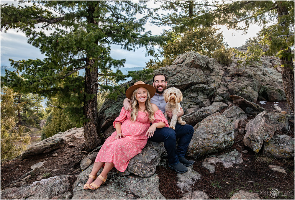 Married couple expecting a baby sit on large boulders in the woods with the cute labradoodle dog at their Colorado maternity pictures