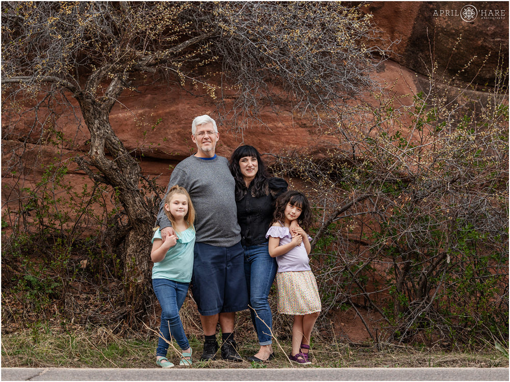 A family of 4 with two daughters pose in front of the trees just barely about to start blossoming at Red Rocks in Colorado