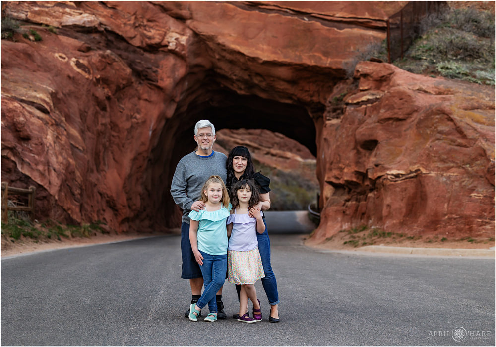 Cute family of four with two daughters pose in front of the famous drive through red rocks arch near the Amphitheater in Colorado