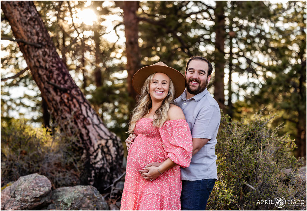 Mom to be wearing a wide brimmed hat and a cute pink off the shoulder sundress poses with her husband at their outdoor maternity photography session in the woods of Colorado