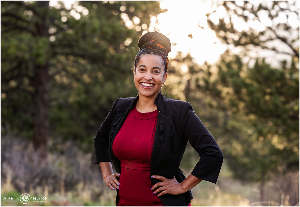 Beautiful woman with a big smile wearing a large bun on top of her head and a maroon colored dress and 3/4 sleeve black jacket for her professional headshot portrait in a forest setting at Shanahan Ridge Trail in Boulder