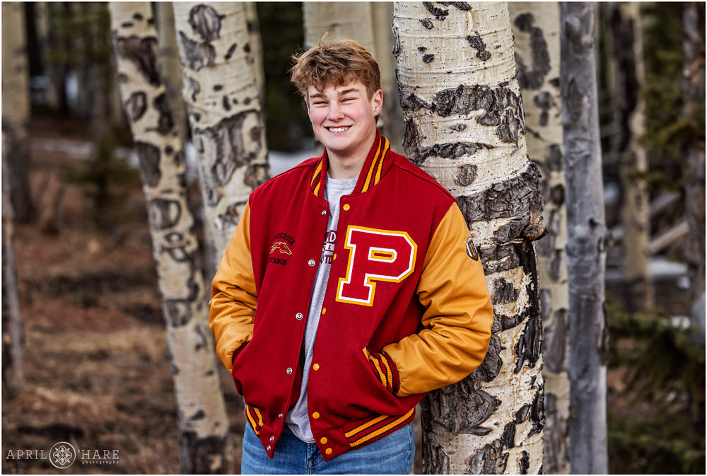 High school senior boy leans up against an aspen tree with red and yellow letter jacket in Evergreen Colorado for his senior portrait session