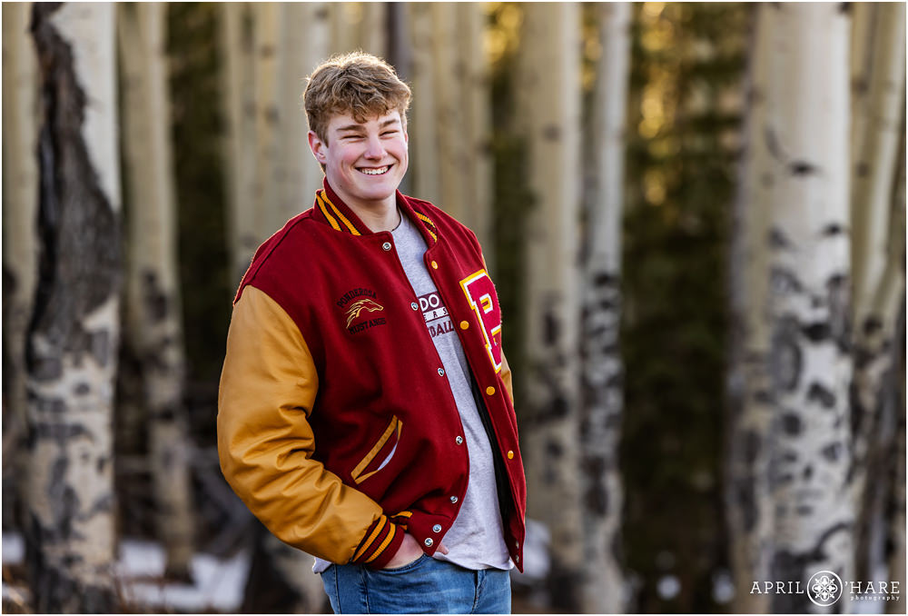 High school senior football player wearing his Ponderosa High School Letter jacket smiling in an Aspen Tree forest in Evergreen Colorado